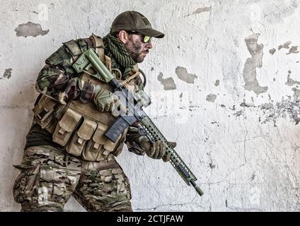 Armeesoldat, Spezialeinsatzkräfte Schütze in Tarnuniform, Lastenträger, Brille und Kappe stehen in der Nähe von abgebrochen Wand mit Service-Gewehr in den Händen. Kommandokämpfer, Kämpfer im Kriegsgebiet Stockfoto