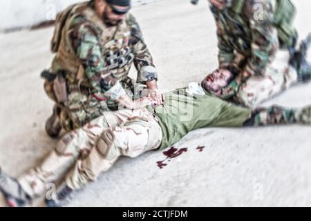 Soldaten, die versuchen, die Blutung des verwundeten Kameraden zu stoppen, der auf dem Boden lag, in paine litt und schrie. Kommandokämpfer, der mit den Händen auf die blutige Wunde auf den Magen der Freunde drückt, die Notfallversorgung abgebend Stockfoto