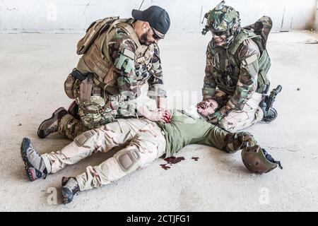 Soldaten, die versuchen, die Blutung des verwundeten Kameraden zu stoppen, der auf dem Boden lag, in paine litt und schrie. Kommandokämpfer, der mit den Händen auf die blutige Wunde auf den Magen der Freunde drückt, die Notfallversorgung abgebend Stockfoto