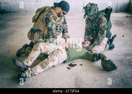 Soldaten, die versuchen, die Blutung des verwundeten Kameraden zu stoppen, der auf dem Boden lag, in paine litt und schrie. Kommandokämpfer, der mit den Händen auf die blutige Wunde auf den Magen der Freunde drückt, die Notfallversorgung abgebend Stockfoto