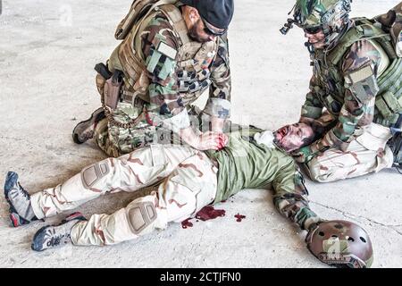 Soldaten, die versuchen, die Blutung des verwundeten Kameraden zu stoppen, der auf dem Boden lag, in paine litt und schrie. Kommandokämpfer, der mit den Händen auf die blutige Wunde auf den Magen der Freunde drückt, die Notfallversorgung abgebend Stockfoto