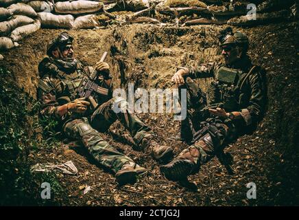Lächelnder US-Armeesoldat, Robbenjäger, moderner Kämpfer in Kampfuniform, Plattenträger, ballistische Brille und Kampfhelm ruhen nach dem Kampf, sitzen in Graben in der Nacht und rauchen Zigarette Stockfoto
