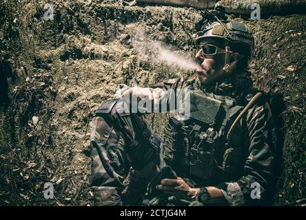 Lächelnder US-Armeesoldat, Robbenjäger, moderner Kämpfer in Kampfuniform, Plattenträger, ballistische Brille und Kampfhelm ruhen nach dem Kampf, sitzen in Graben in der Nacht und rauchen Zigarette Stockfoto