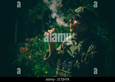 Kommandosoldat in Tarnuniform, ballistische Brille und bonnie, inhalieren Zigarettenrauch, Rauchen Tabak oder Marihuana Joint in Wald oder Dschungel in der Nacht. Armee Infanterist entspannend nach dem Kampf Stockfoto