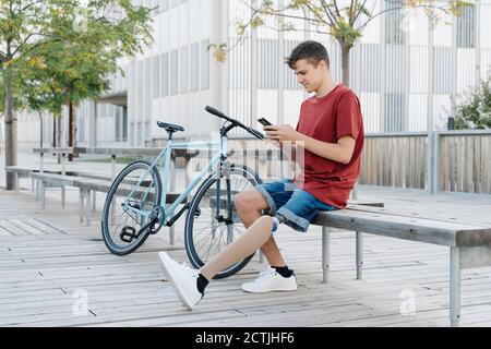 Teen männlich mit Bein Prothese sitzt auf Bank in der Nähe Fahrrad Und das Surfen auf dem Smartphone Stockfoto