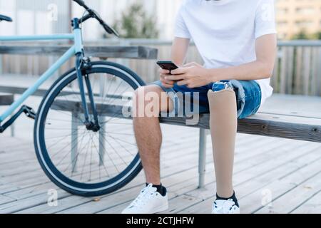 Männlicher Radfahrer mit Beinprothese sitzt auf der Bank und mit Mobiltelefon Stockfoto