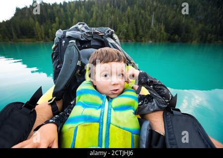 Junge trägt PFD ruht gegen Rucksack auf aufblasbarem Boot auf Blauer See Stockfoto