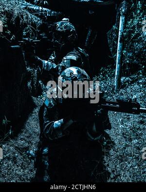 Zwei Soldaten der Armee, Navy siegelt Team Schützen tragen Kampfuniform, Körperpanzerung und Helm, zielende Service Gewehre, die sich gegenseitig während der Bewegung in Graben. Infanteristen auf dem Schlachtfeld bei Nacht Stockfoto