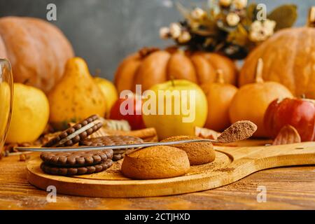 Schokoladenkekse und Herbsternte auf Holztisch. Reife Kürbisse, Äpfel und Kaki auf dem Hintergrund. Löffel mit Gewürzen oder Schokoladenstückchen. Das Konzept von Weihnachten, Thanksgiving und Halloween. Stockfoto