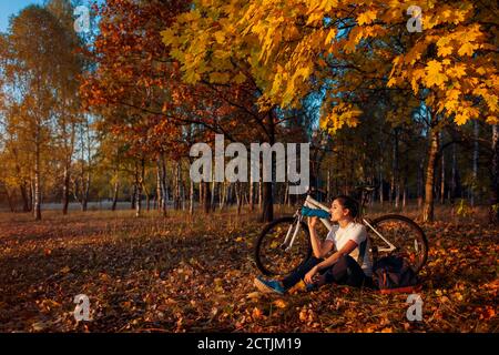 Junge Frau trinkt Wasser nach dem Training auf dem Fahrrad. Gesunder Lebensstil. Fahrrad fahren im Herbstwald. Stockfoto