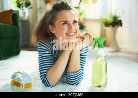 Lächelnd moderne Frau im gestreiften T-Shirt und weißen Hosen mit Sprühflasche mit umweltfreundlichen Reinigungsmitteln und gelber Bürste Auf weißem Teppich liegend Stockfoto