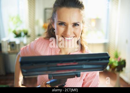 Lächelnde junge Frau in Bluse und weißen Hosen mit Staubsauger Bürste in der modernen Haus in sonnigen Tag. Stockfoto