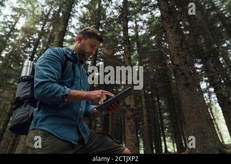 Mann überprüft GPS über digitales Tablet, während er gegen Bäume steht Im Wald Stockfoto