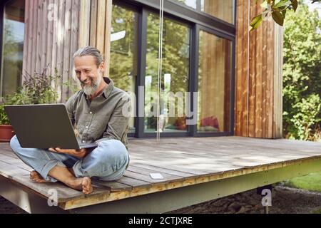 Bärtiger reifer Mann mit Laptop, während er vor dem winzigen Haus sitzt Stockfoto