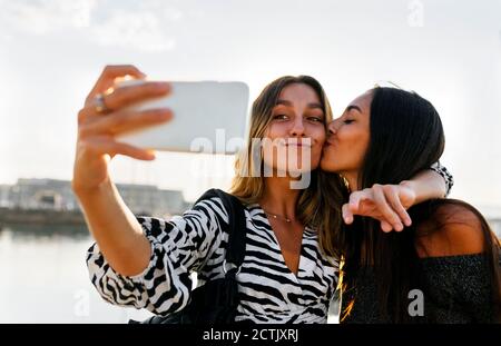 Junge Frau küsst Freund während Selfie gegen klaren Himmel Stockfoto