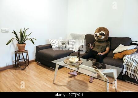 Mann trägt Teddybärmaske und schaut beim Entspannen fern Sofa im Wohnzimmer Stockfoto