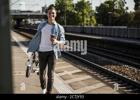 Lächelnder junger Mann, der einen Roller trägt, während er auf der Eisenbahn läuft Bahnsteig Stockfoto