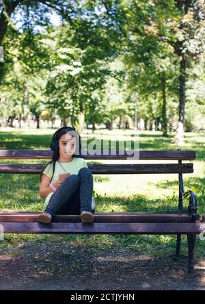 Nettes Mädchen mit Kopfhörer sitzen auf Bank im Park Stockfoto