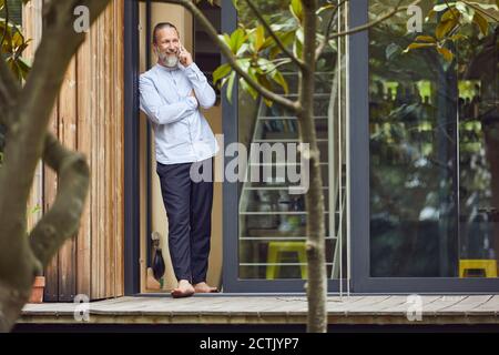 Bärtiger reifer Mann, der über Smartphone spricht, während er ansteht Eingang von in winzigen Haus Stockfoto
