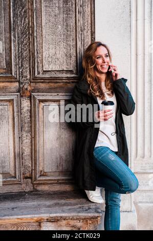 Schöne Frau, die am Telefon telefoniert und Kaffee gegen die Holzwand hält Im Park Stockfoto