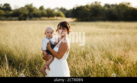 Braut trägt Baby Junge, während auf dem Feld während des Sonnenuntergangs stehen Stockfoto