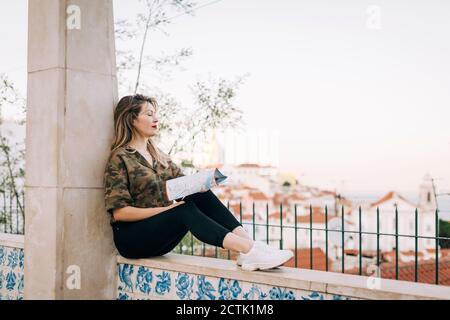 Junge Frau mit Karte sitzt auf Stützmauer gegen klar Himmel Stockfoto