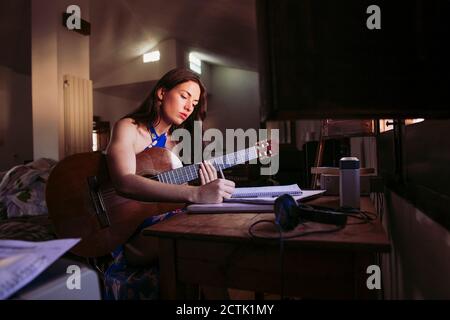 Junge Frau, die im Buch schreibt, während sie zu Hause Gitarre übt Stockfoto