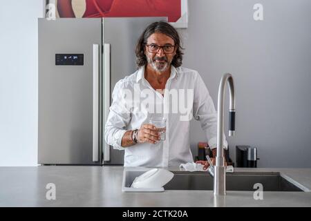 Lächelnder Mann, der Wasser trinkt, während er am Waschbecken in der Küche steht Stockfoto