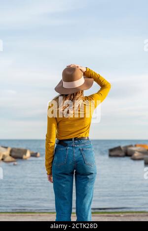 Junge Frau mit erhobenen Armen trägt Sonnenhut gegen Meer Stockfoto