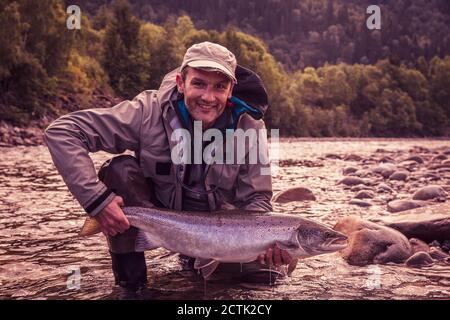 Fliegenfischer lächelnd, während gefangener Lachsfisch im Fluss gehalten wird Stockfoto