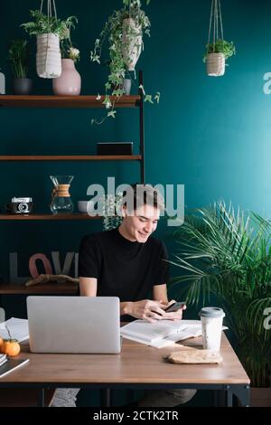 Lächelnder junger Mann mit Smartphone, während er mit Büchern sitzt Und Laptop am Tisch Stockfoto