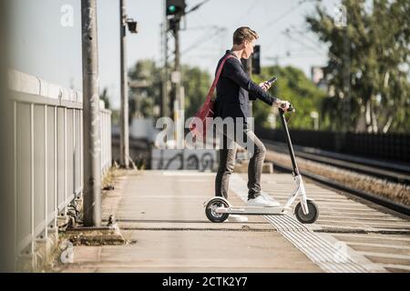 Mann mit Smartphone während der Wartezeit mit Push Scooter an Bahnhofsplattform Stockfoto