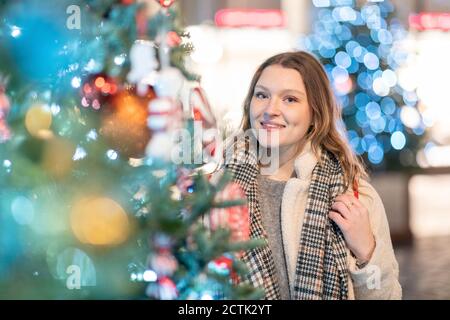 Lächelnd schöne Frau, die in der Stadt beim beleuchteten Weihnachtsbaum steht Nachts Stockfoto