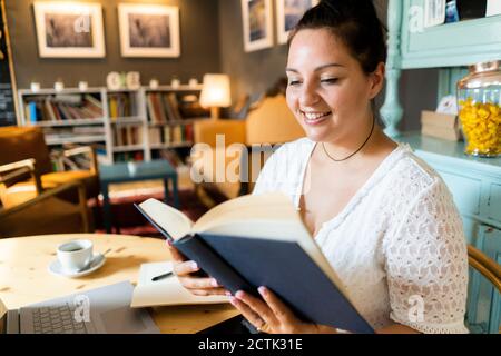 Lächelnde üppige Frau liest Buch, während sie an einem Tisch sitzt Restaurant Stockfoto