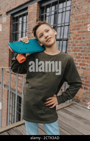 Nachdenklicher Junge posiert mit blauem Skateboard, während er gegen Gebäude steht Stockfoto