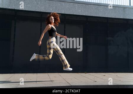 Lächelnde Frau mit zerzausten Haaren springt auf Bürgersteig in der Stadt Stockfoto