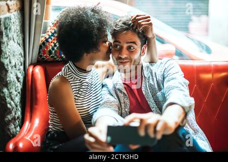 Mann, der Selfie nimmt, während Freundin ihn küsst, während er auf sitzt Sofa im Café Stockfoto