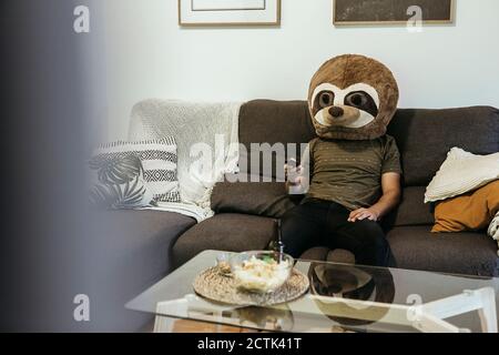 Mittelerwachsener Mann mit Teddybärmaske beim Fernsehen Sitzen im Wohnzimmer Stockfoto