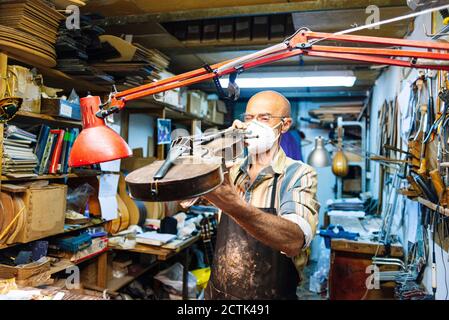 Geigenbauer mit Gesichtsmaske zur Prüfung der Violine in der Werkstatt Stockfoto