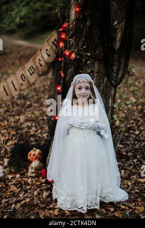 Kleines Mädchen stehend, während das Kostüm der Leiche Braut gegen den Baum trägt Im Wald Stockfoto