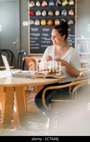 Lächelnde üppige Frau hält Kaffeetasse während des Studiums über Laptop Im Restaurant Stockfoto