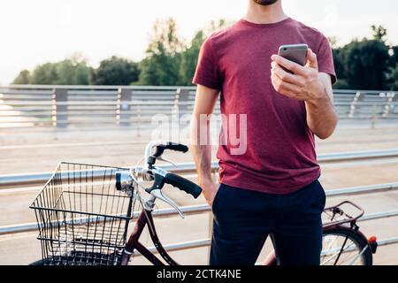 Nahaufnahme eines Mannes mit Smartphone, während er mit dem Fahrrad steht Auf der Brücke bei Sonnenuntergang Stockfoto