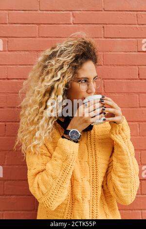 Junge Frau trinkt Kaffee aus Einweg-Tasse beim Blick weg Gegen rote Backsteinmauer Stockfoto