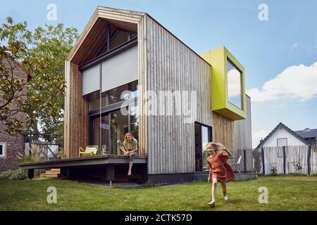 Mutter schaut auf fröhliche Tochter, die im Hof läuft, während sie sitzt Vor einem winzigen Haus Stockfoto