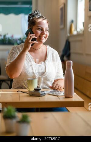 Üppige junge Frau, die über das Mobiltelefon spricht, während sie ansitzt Tisch im Restaurant Stockfoto