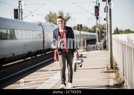 Lächelnder junger Mann, der einen Roller trägt, während er auf der Eisenbahn läuft Bahnsteig Stockfoto