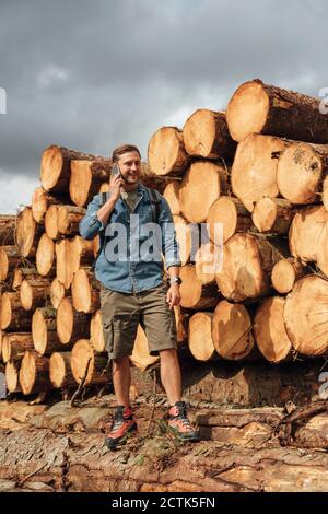 Mann mit mittlerem Erwachsenenalter, der im Stehen über ein Smartphone spricht Holzstapel im Wald Stockfoto