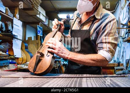 Handwerker mit Gesichtsmaske Prüfung Saite der Geige auf Werkbank In der Werkstatt Stockfoto