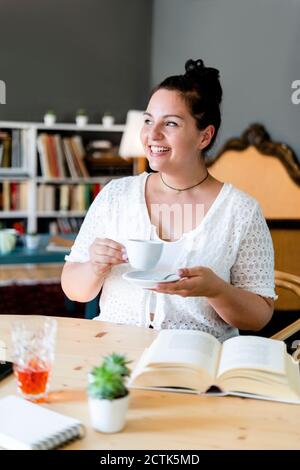 Glückliche junge Frau mit Buch auf dem Tisch mit Kaffeetasse Beim Sitzen im Restaurant Stockfoto