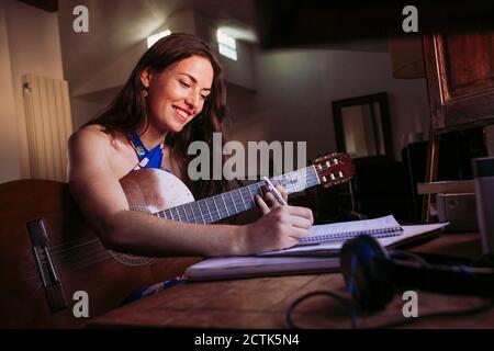 Lächelnde junge Frau, die im Buch schreibt, während sie Gitarre übt Zu Hause Stockfoto